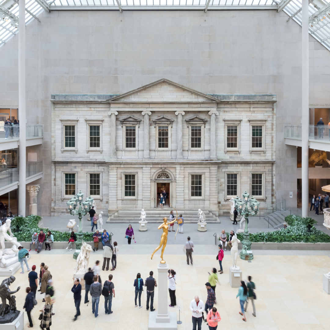 Photo of the American Wing gallery at The Metropolitan museum of art: an indoor courtyard with sky lights with the facade of a bank vault on one wall with people roaming around the courtyard.
