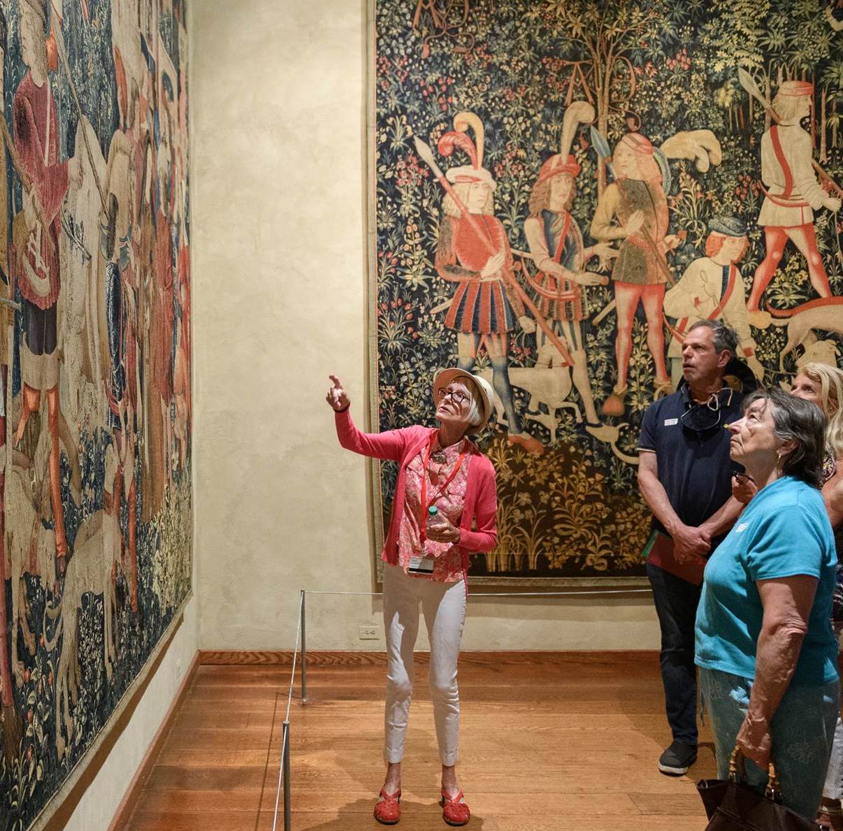 An older woman in a pink cardigan leads a group of visitors and points to a hanging medieval tapestry at The Met Cloisters.