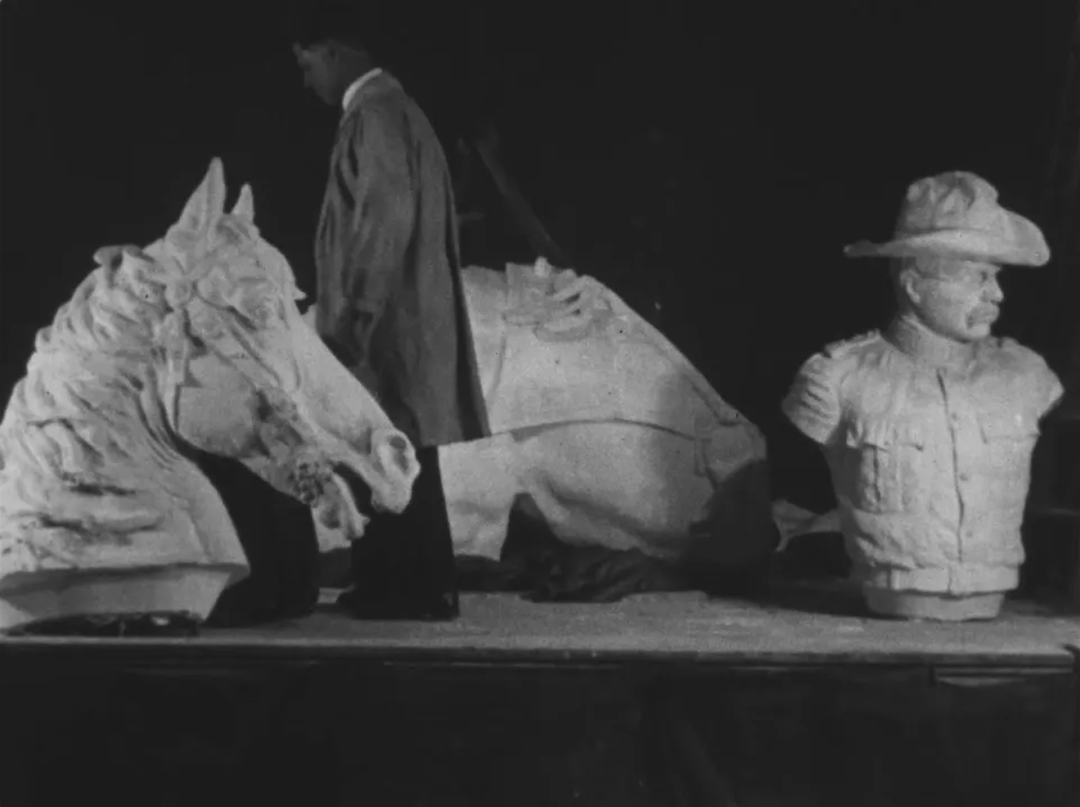 Several plaster mold models of a horse's head and a man's torso on a platform, while a human figure walks around to assess the fragmented sculptures.