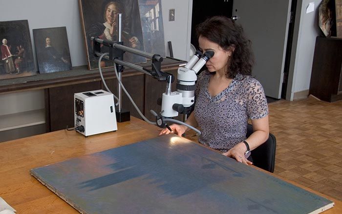 Conservator Isabelle Duvernois examining a Monet landscape through a microscope in the Paintings Conservation studio. Unframed paintings are leaning against a wall on a ledge in the background.